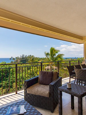 A spacious balcony with outdoor seating and dining area overlooks a lush landscape and ocean view, creating a relaxing atmosphere.